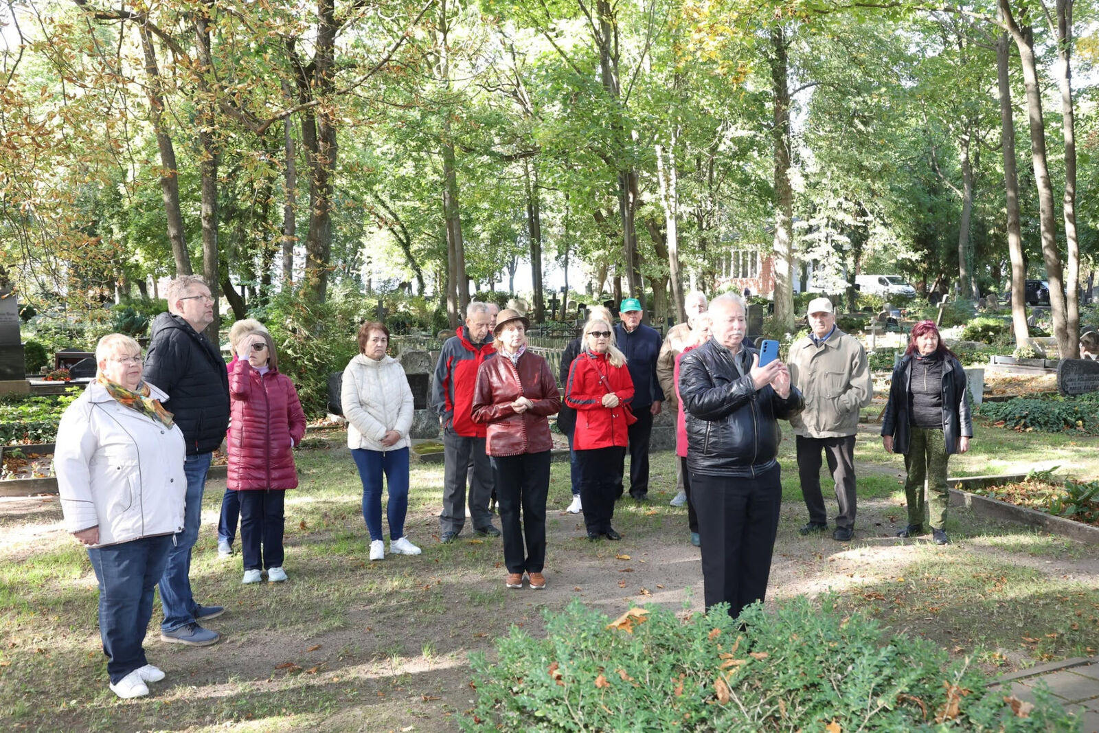 Dalībnieki stāv parkā starp kokiem un klausās runātājā