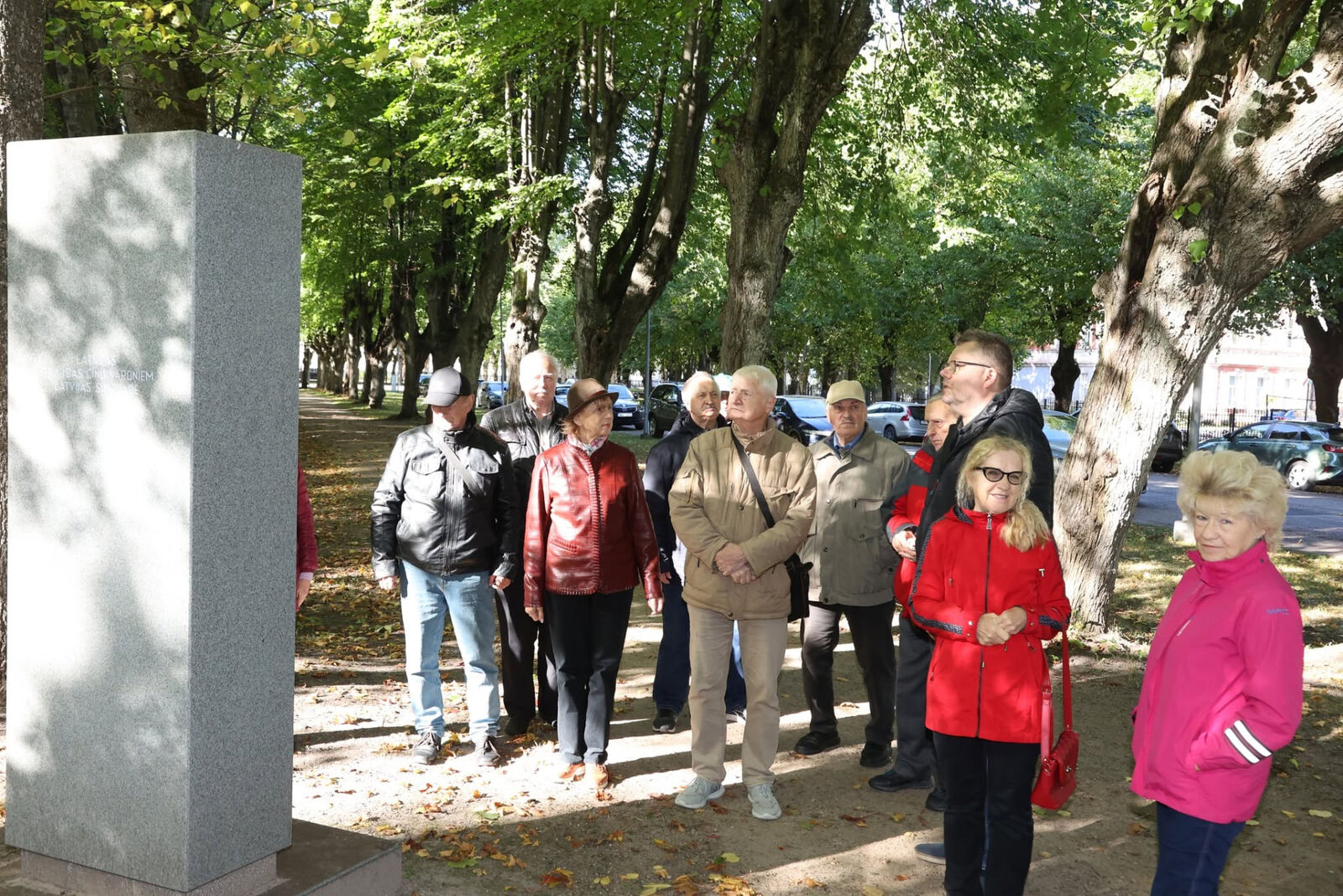 Dalībnieki koku alejā pie gaiša akmens piemiņas zīmes